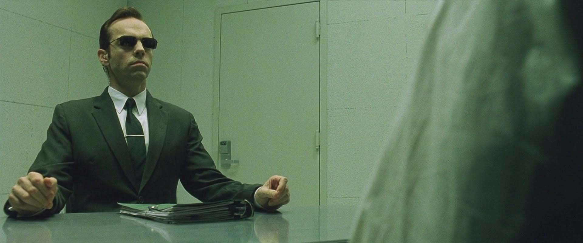 Agent Smith, interpretato da Hugo Weaving, al tavolo durante l’interrogatorio di Neo in The Matrix (1999).